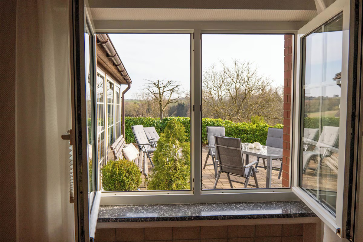 Geöffnetes Fenster mit Blick auf die Terrasse mit Liegestühlen und die Felder dahinter
