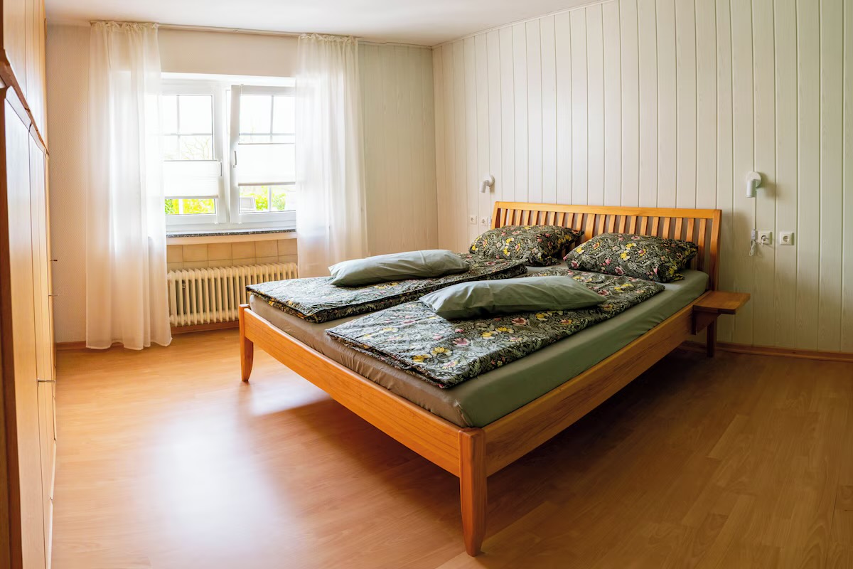Schlafzimmer mit Doppelbett aus Holz, Fenster mit Vorhängen und hellem Dielenboden