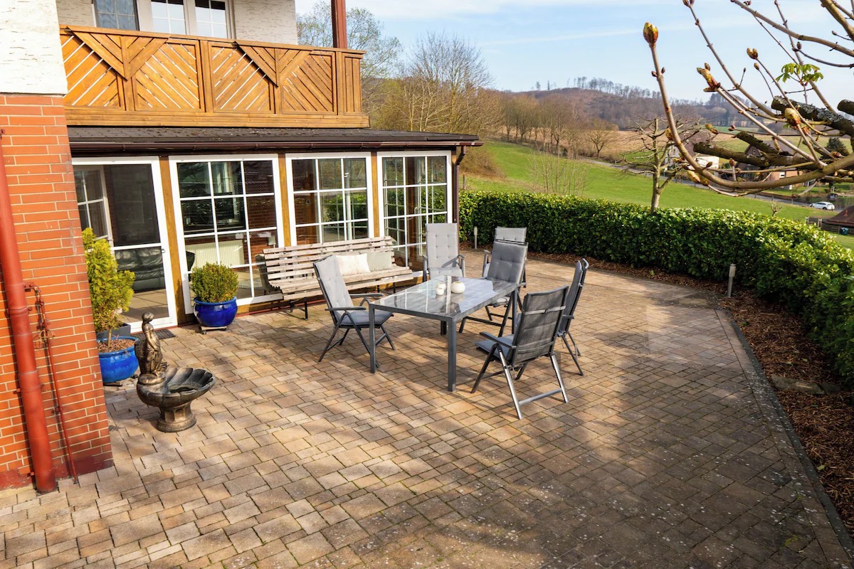 Große gepflasterte Terrasse mit Tisch, Klappstühlen und Bank vor Hecke, dahinter Wiesen und Hügel