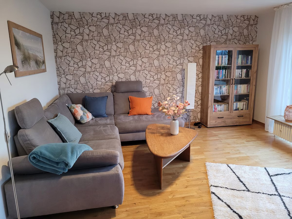 Große Eckcouch mit Kissen und Decke vor einer Steinoptik-Wand, daneben ein gut gefüllter Bücherschrank